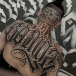 A person's upper back and neck featuring a large, intricate black and grey script tattoo.