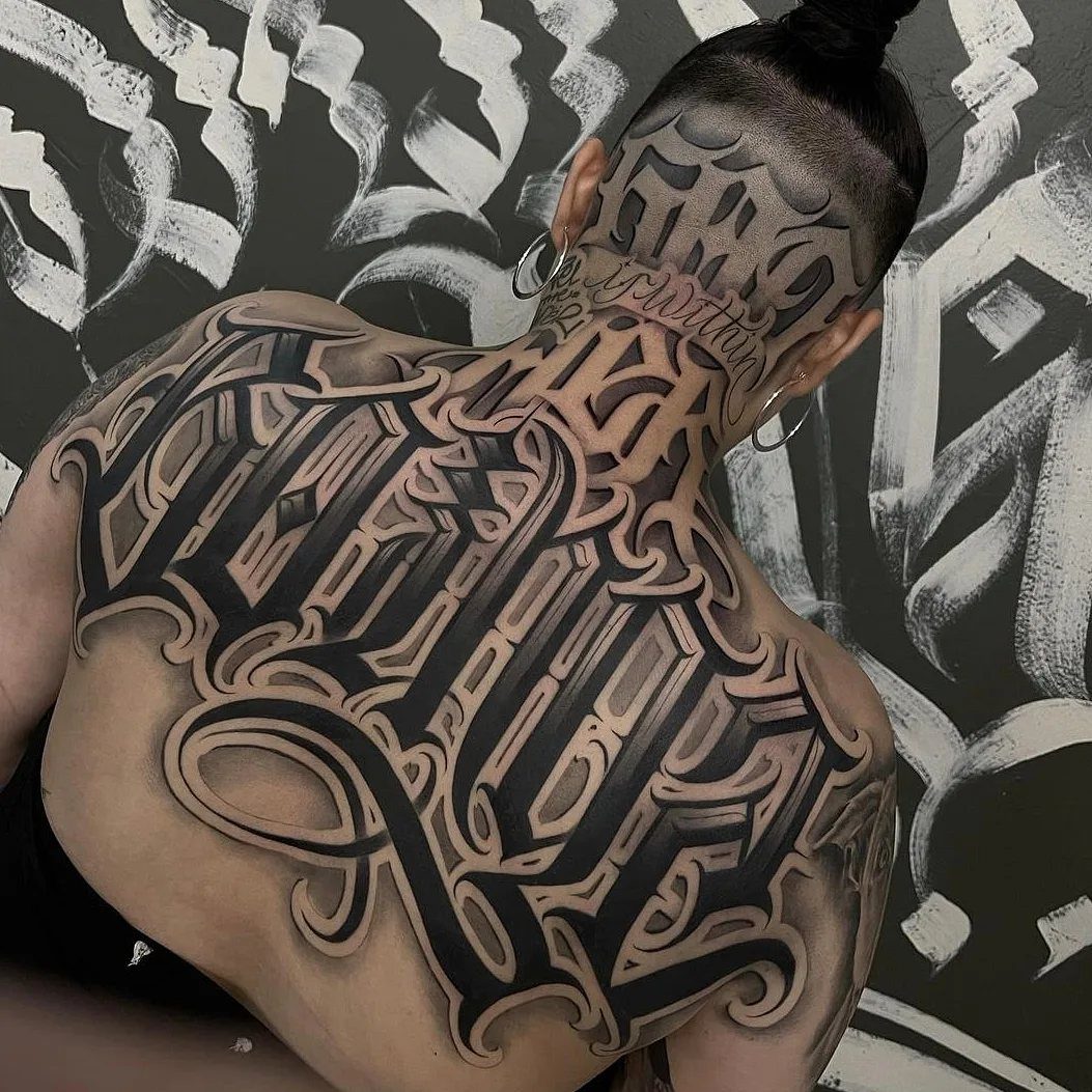 A person's upper back and neck featuring a large, intricate black and grey script tattoo.