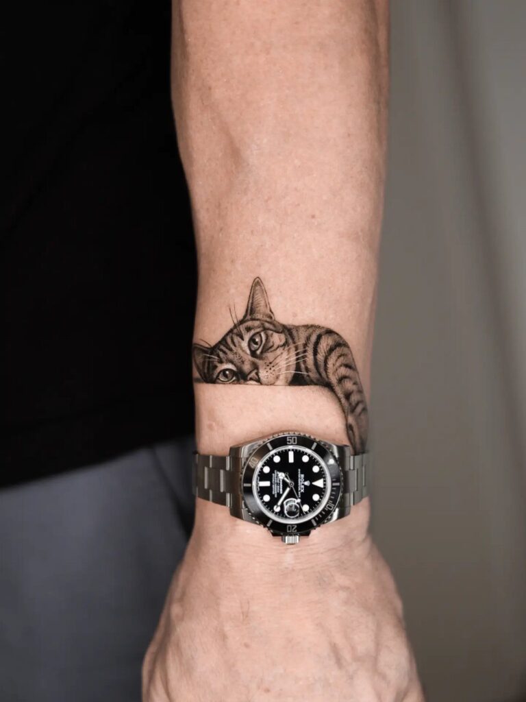 A realistic black and grey tabby cat tattoo peeking over a person's wrist and watch.