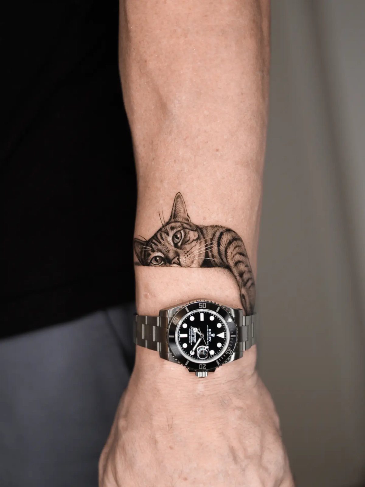 A realistic black and grey tabby cat tattoo peeking over a person's wrist and watch.