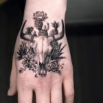 A detailed black and grey cow skull tattoo with cacti on a person's hand.