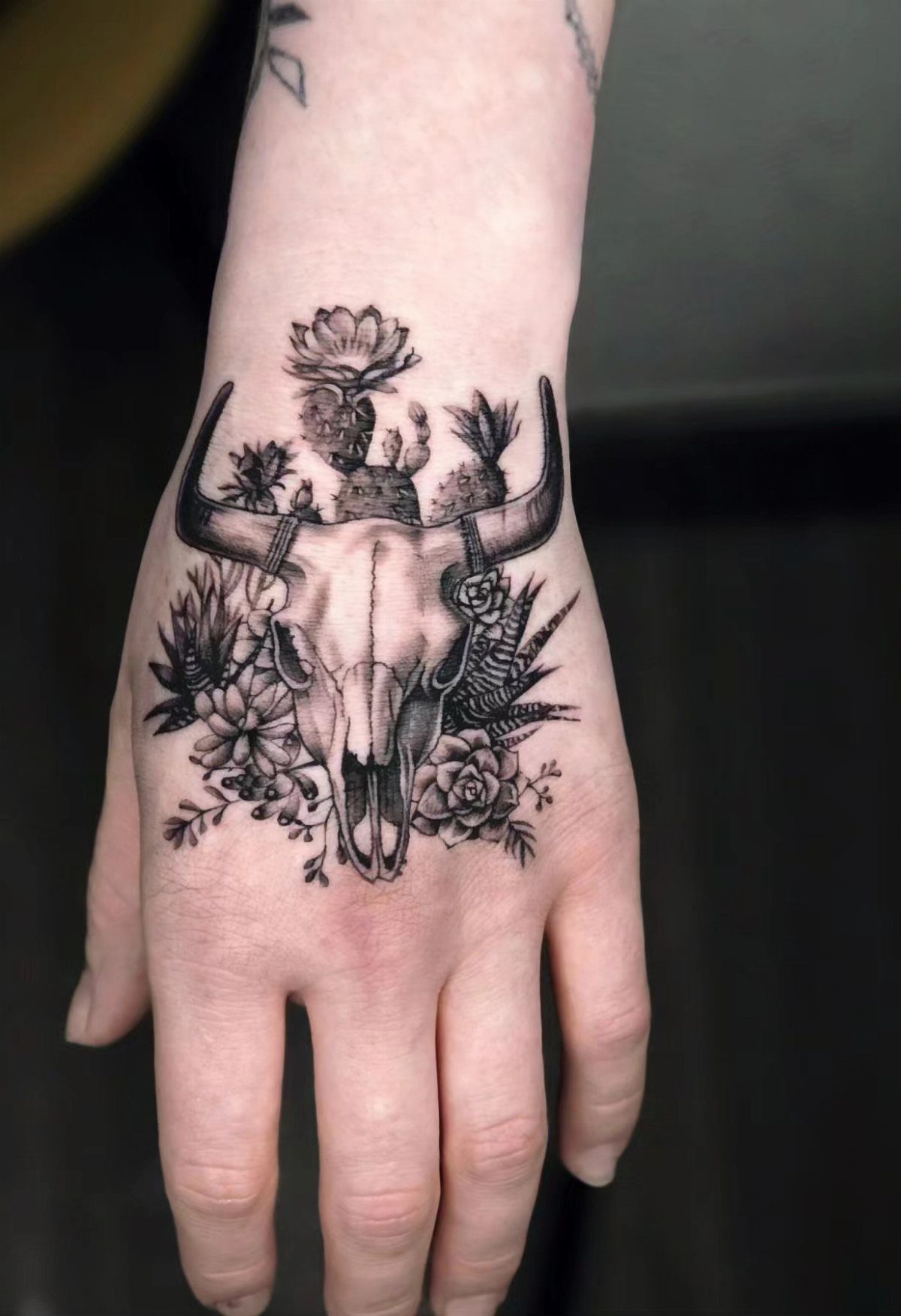 A detailed black and grey cow skull tattoo with cacti on a person's hand.