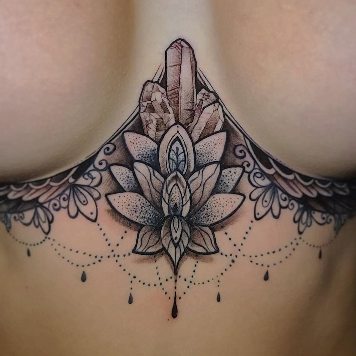 A detailed black and grey lotus and crystal tattoo on a woman's sternum.