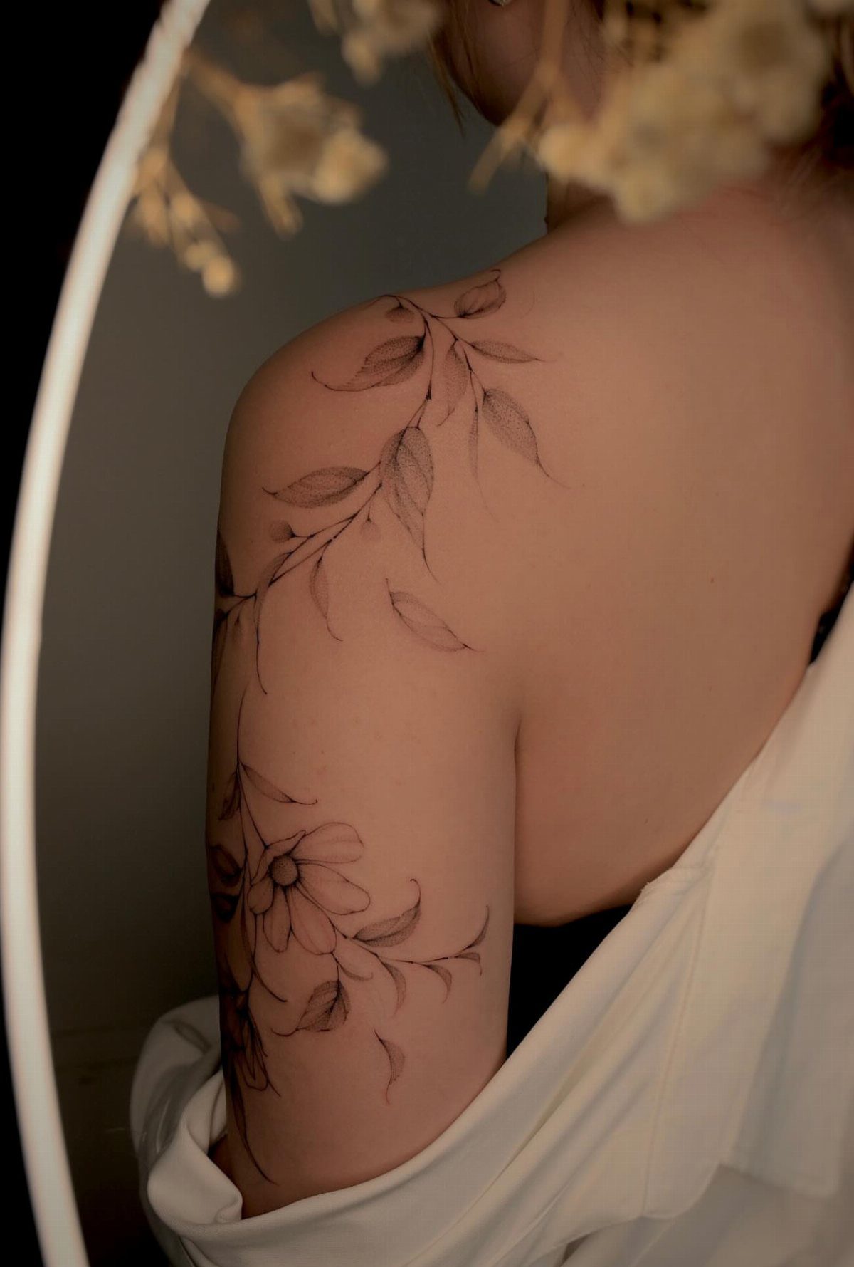 A fine line flower and vine tattoo winding around a person's shoulder and arm.