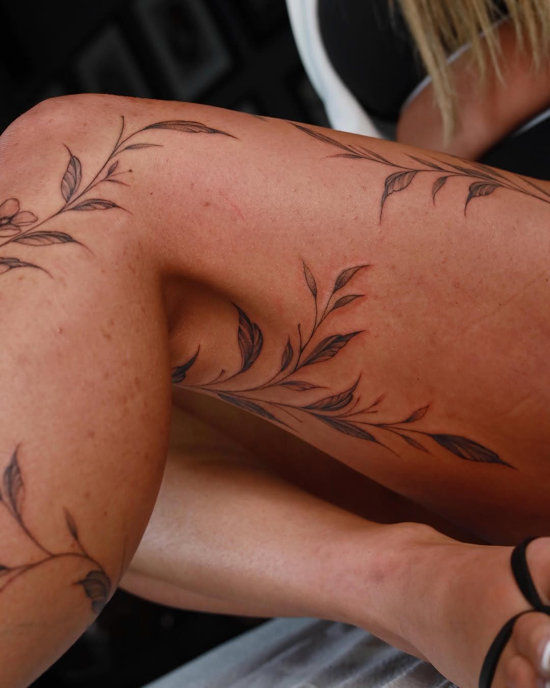 Black ink leaf and vine tattoos winding around a person's freckled leg and thigh.