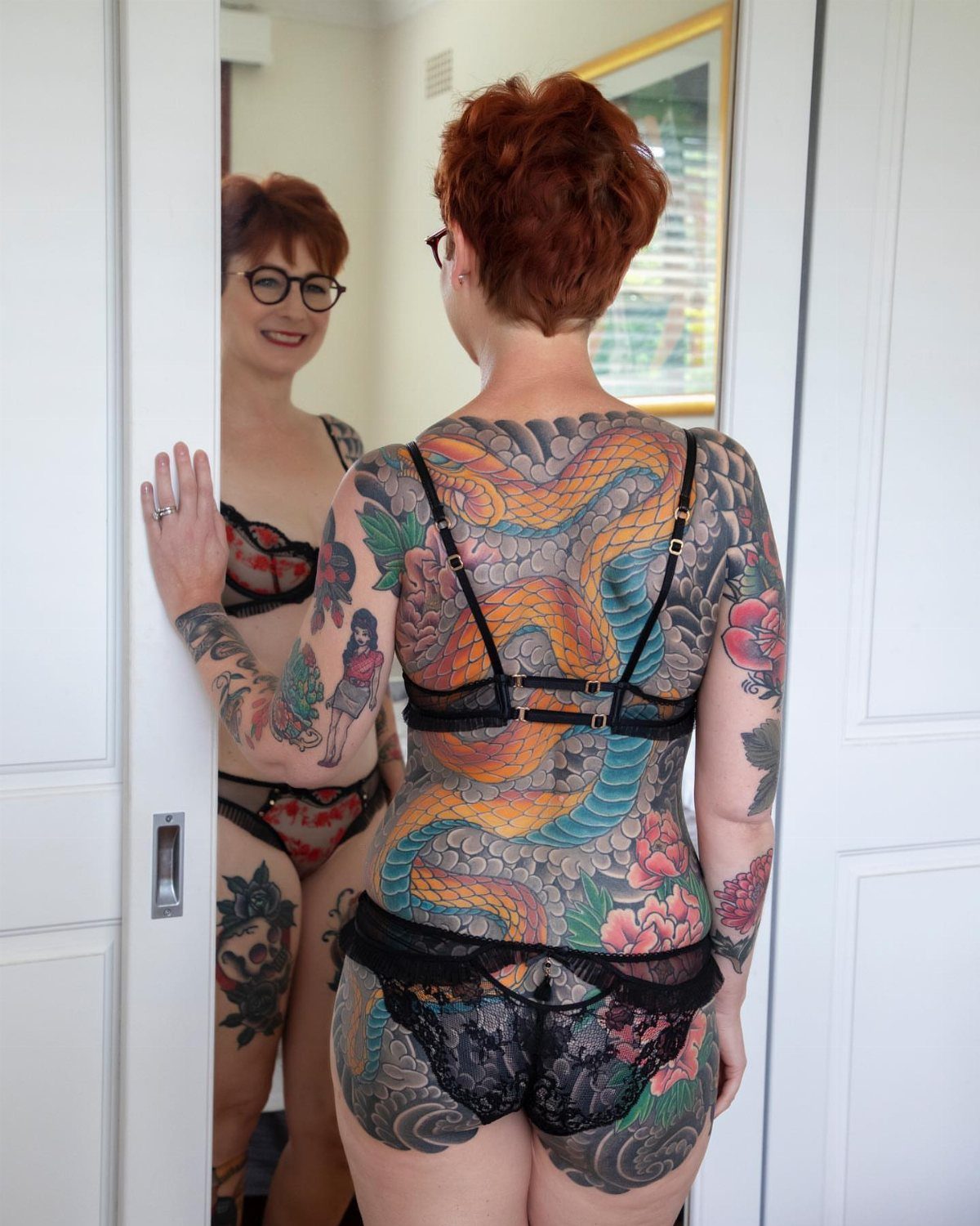 A woman with red hair shows her large, colorful Japanese-style back tattoo in a mirror.