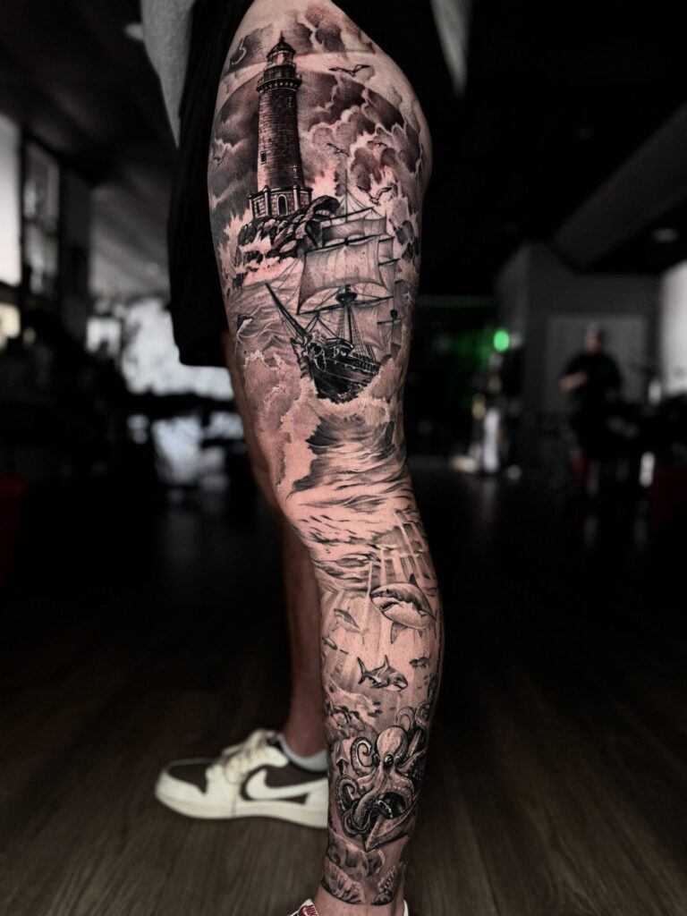 A detailed black and grey tattoo on a leg depicting a lighthouse, ship, and octopus.