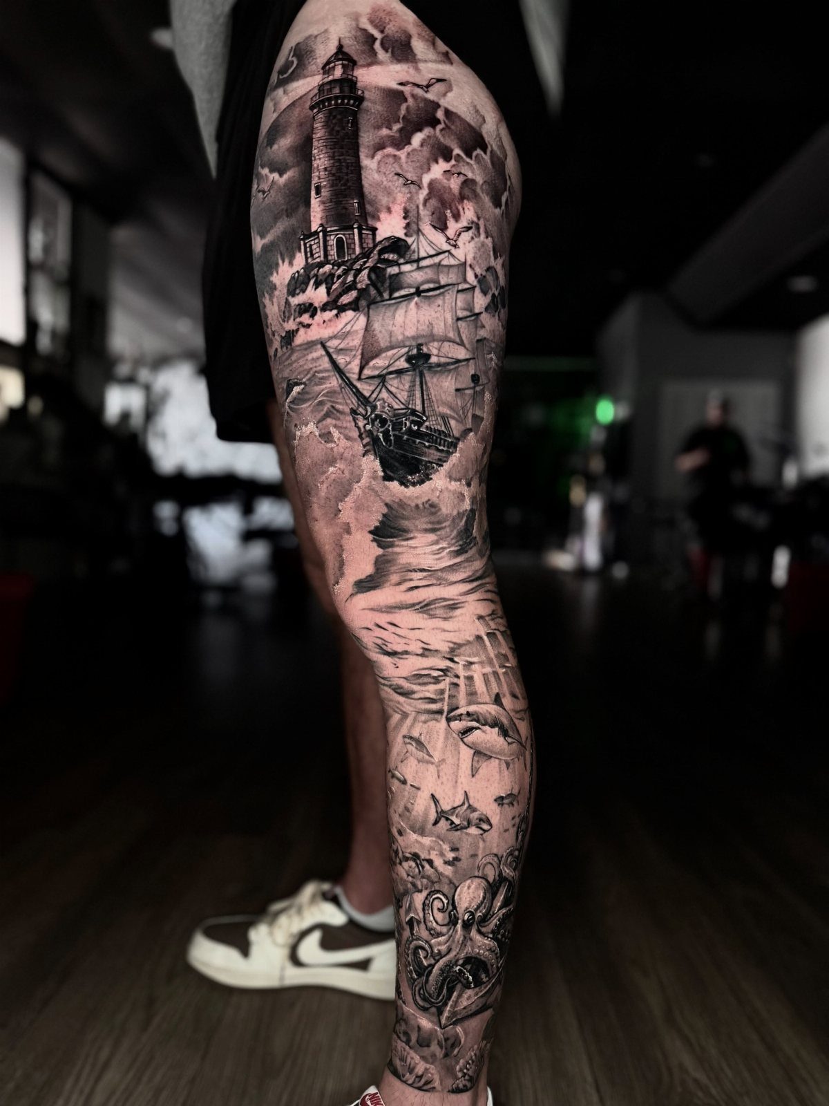 A detailed black and grey tattoo on a leg depicting a lighthouse, ship, and octopus.