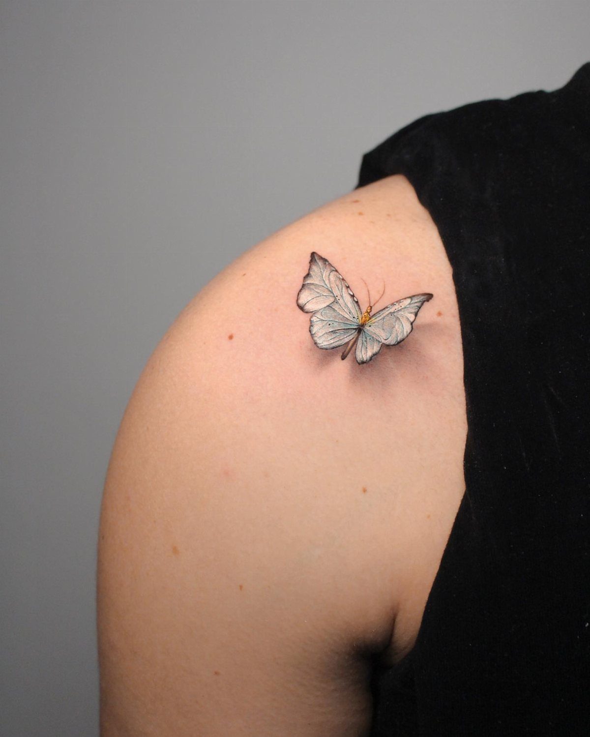 A detailed micro-realistic butterfly tattoo on a person's shoulder against a grey background.