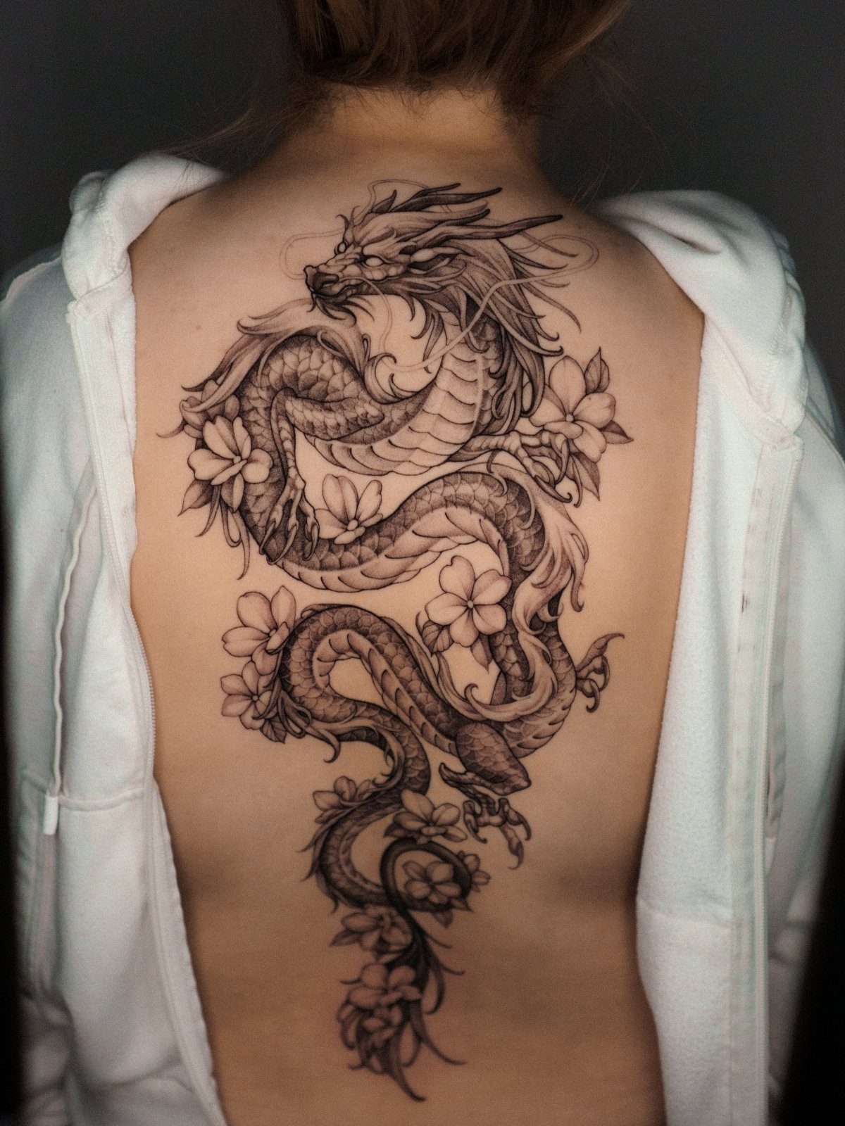 A large black ink dragon tattoo with flowers running down a woman's spine.