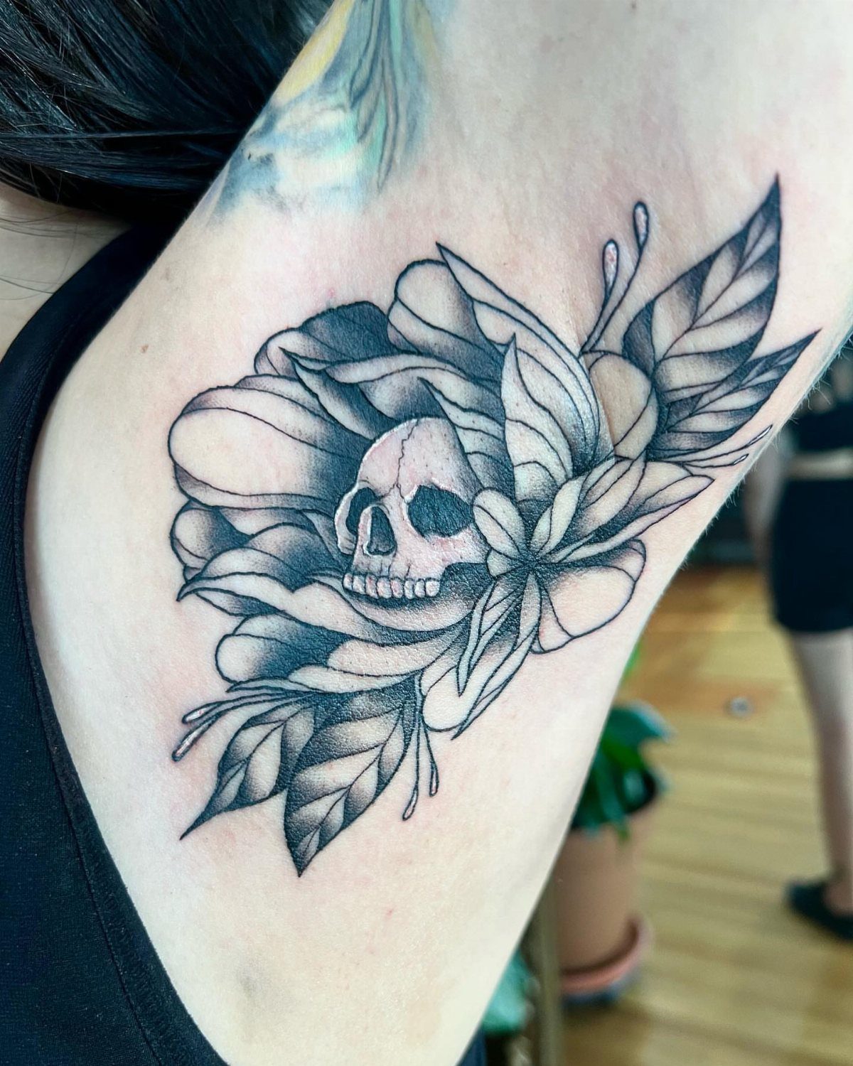 A black and grey floral tattoo featuring a skull centered among large peony petals on a person's ribs.