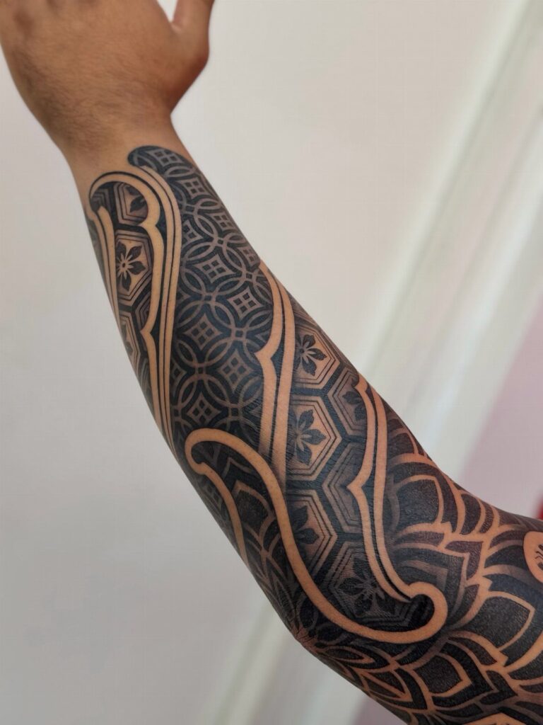 A person's forearm displaying a large, intricate black ink tattoo with complex geometric patterns and flowing lines.